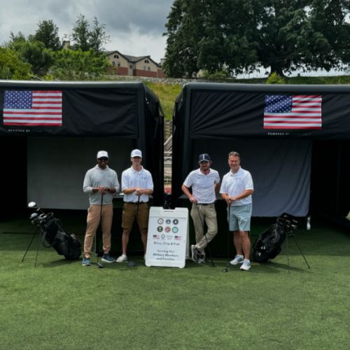 Four men stand with golf clubs in front of two black tents with American flags at a golf event on a sunny course.