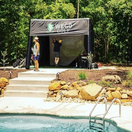 Golf practice tent labeled 'WEGO GOLF' with two people near the entrance, one swinging a club, pool area in the foreground